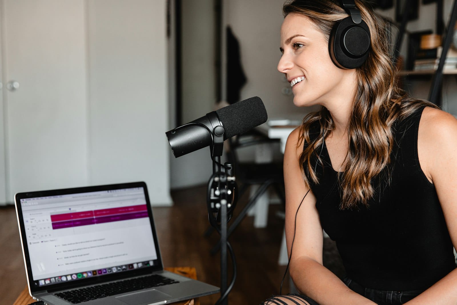 Mikrofon başında kulaklıkla podcast kaydı yapan bir kadın; önünde ses düzenleme programı açık bir dizüstü bilgisayar
